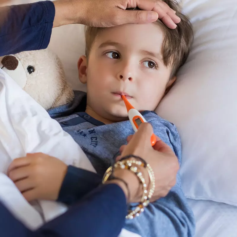 A child in bed with a fever, their parent holding a thermometer in his mouth.