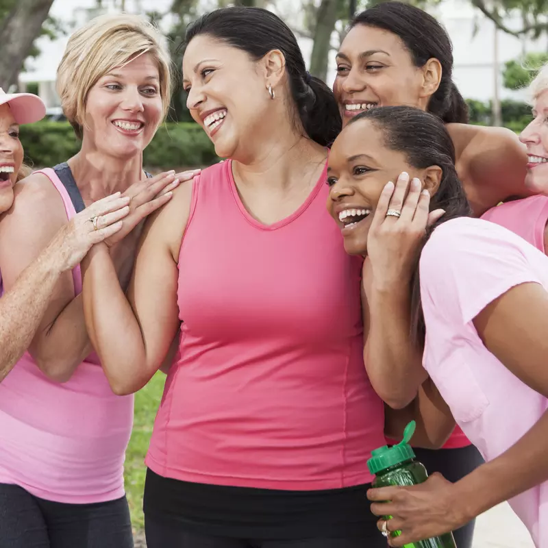 A group of friends, all wearing pink and hugging a woman in the venter, to support a breast cancer survivor.