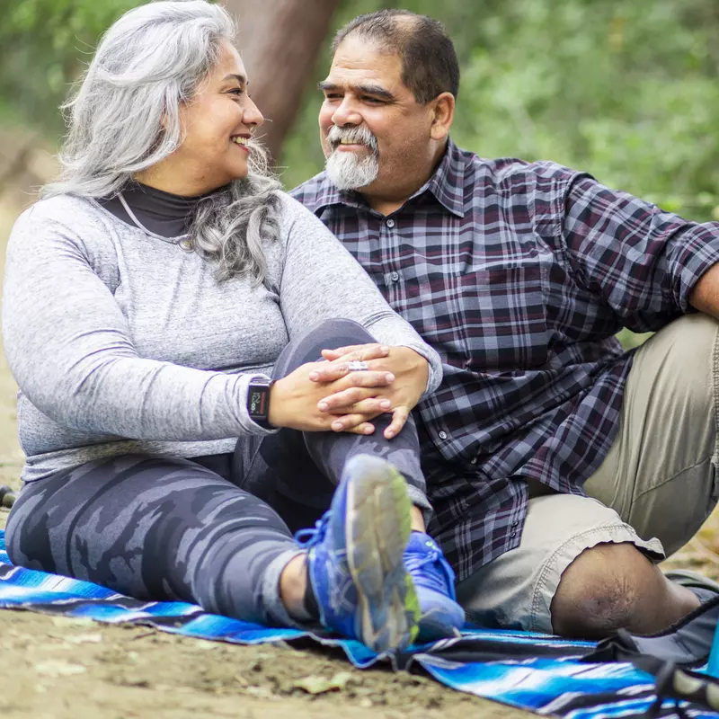 A senior couple is sitting on the ground outdoors enjoying a picnic after a hike.