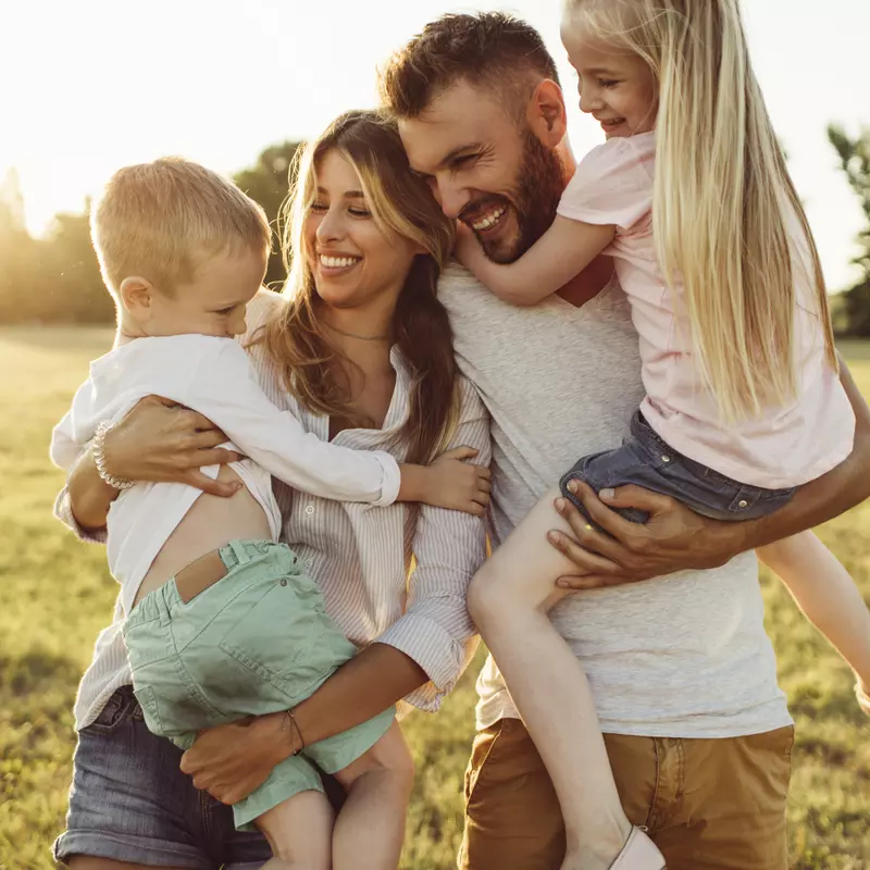 A family of four, the mother holding the son and the father holding the daughter, all laughing and smiling. The sun setting behind them.