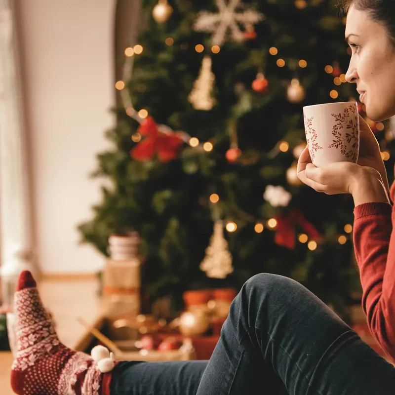 Woman sitting on the ground next to a Christmas tree, drinking something from a mug.