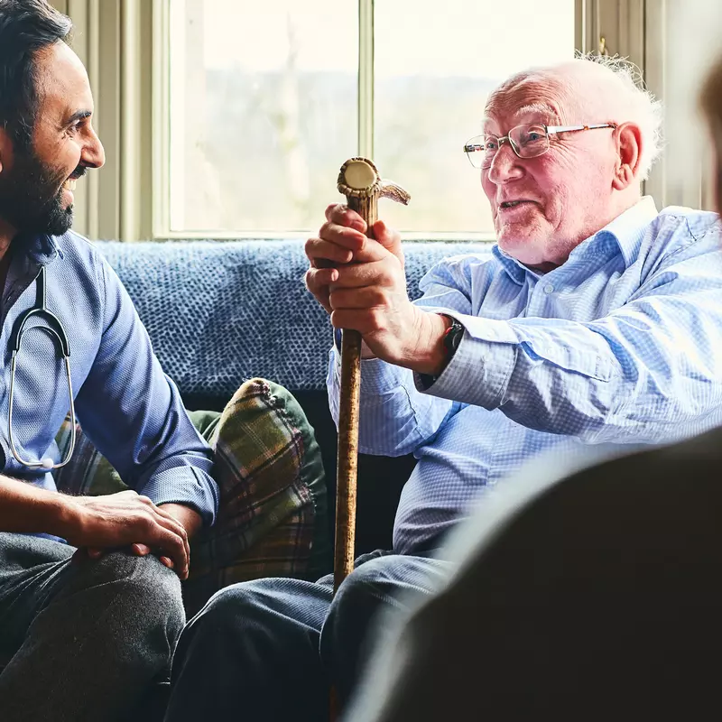 A doctor, sitting on a sofa with a senior man holding a cane, making a home visit.