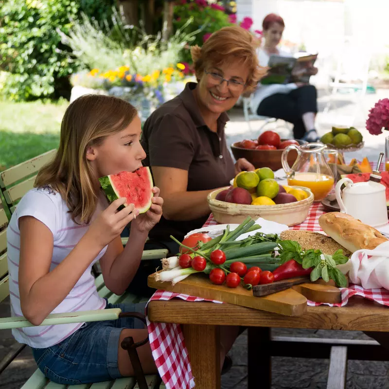 A family of four enjoys a summer picnic, outdoors, sitting at a picnic table enjoying summer foods.