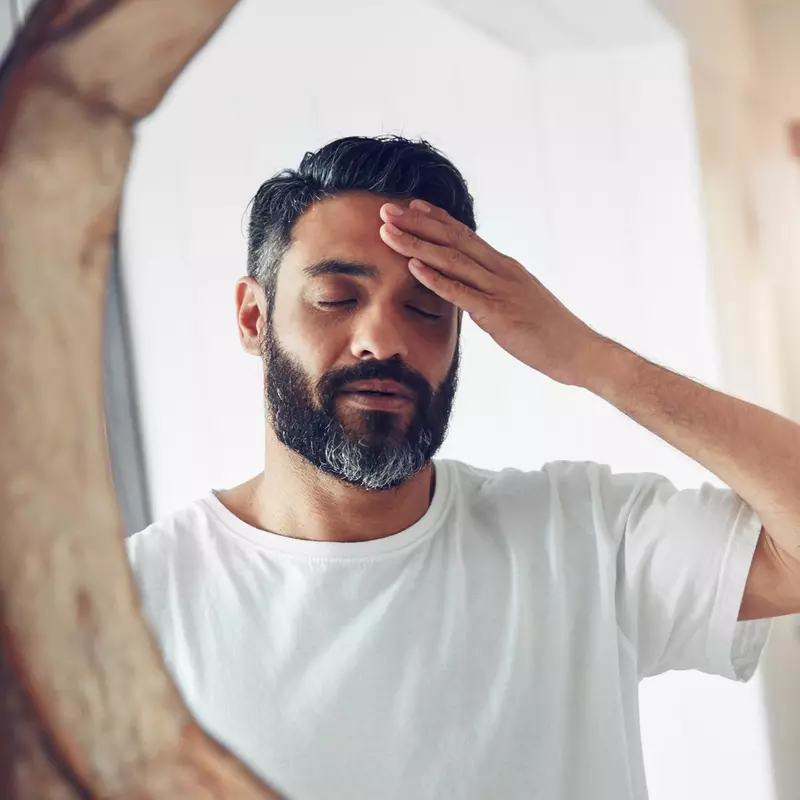 A man is standing in front of a mirror, rubbing his head, stressed out.