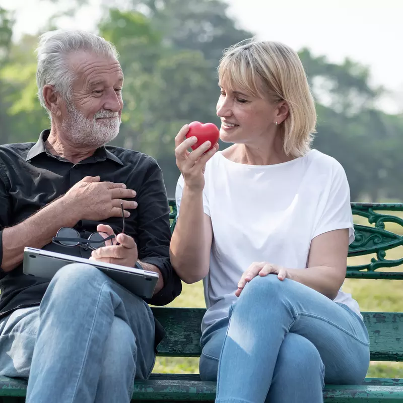 A senior couple sitting on a bench in the park, the woman holding a red heart.
