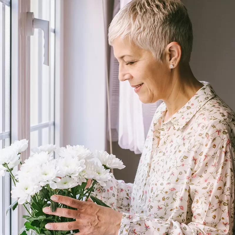 An adult caucasian woman arranges a vase of flowers by the window.