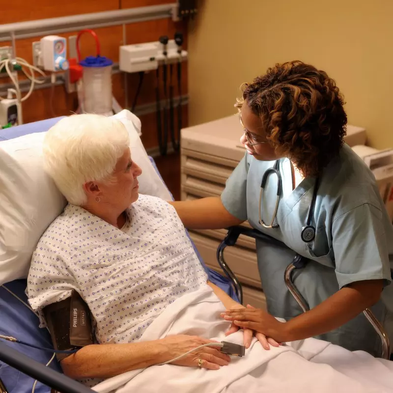 An older woman laying in a hospital bed talking to a nurse.
