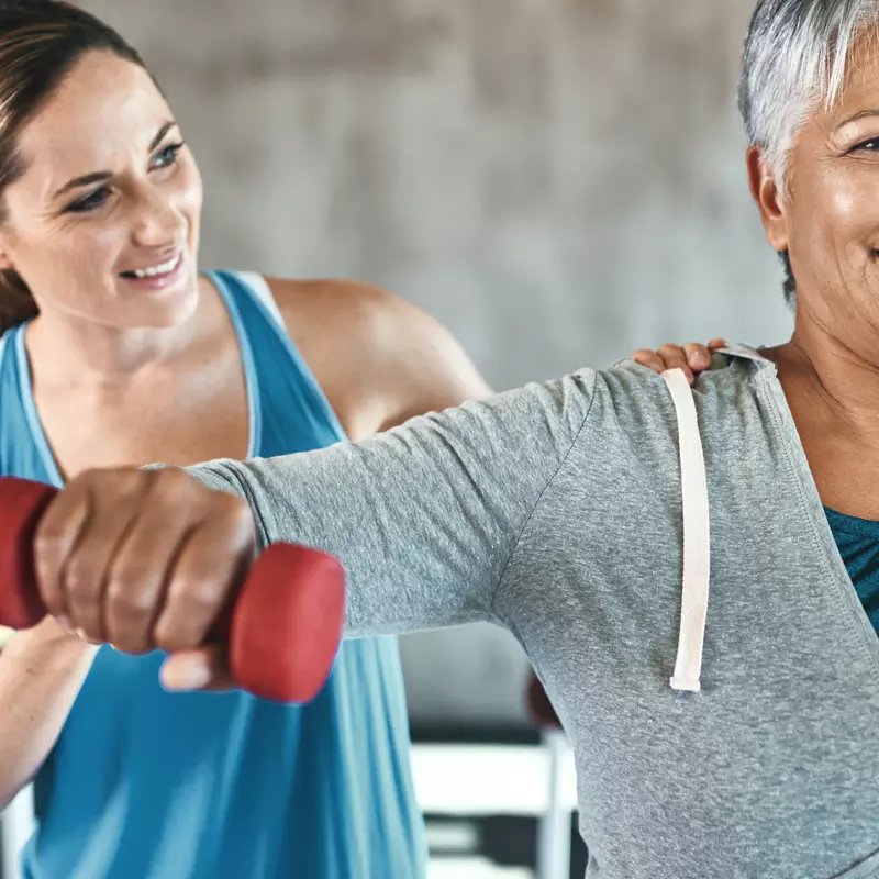 Adult woman with a grey sweatshirt, lifting red weights, with a younger woman in a blue tank top coaching her.