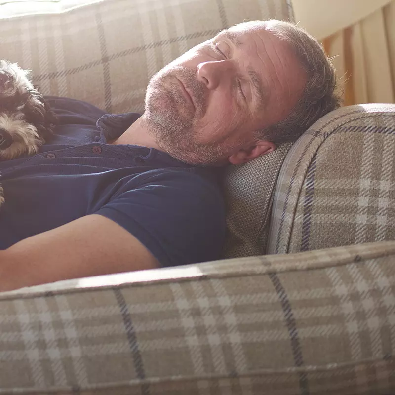 A middle aged man asleep on a patterned sofa with a white and black curly haired dog on asleep on top of him.