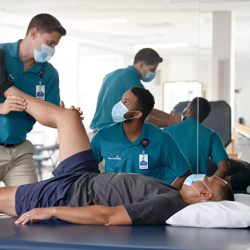 AdventHealth Sports Medicine and Rehab team members helping a patient, who is laying down on a table. His right leg is extended upwards, with a bend in his knee. One of the therapists is holding his leg at the calf and the knee, and looking down at the other therapist who is seated next to him. 