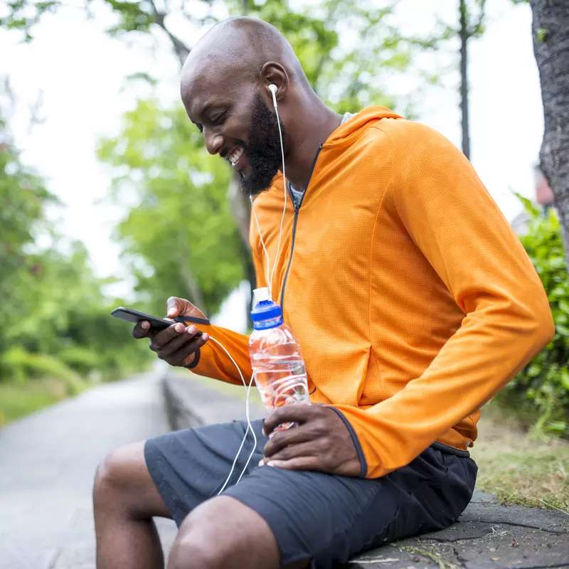 An adult man in an orange hoodie, sitting on a bench outside, looking at his phone and hydrating with a bottle of water.