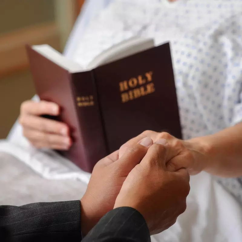 A chaplain and a patient holding hands and reading the bible