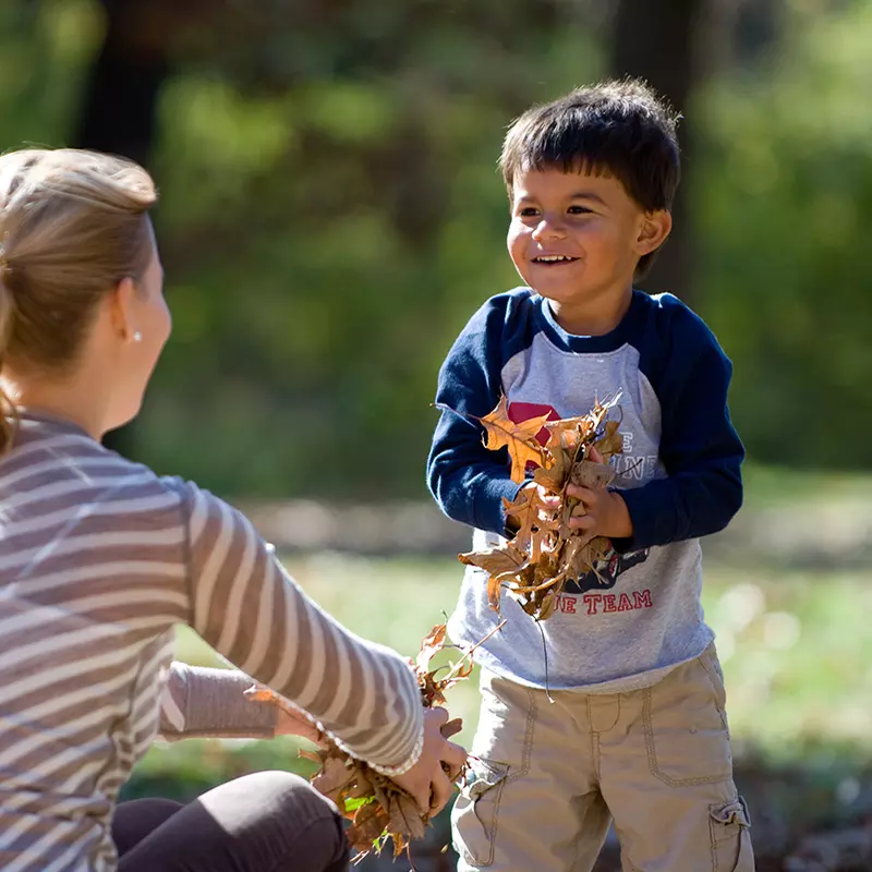 A boy plays happily in the fallen leaves with his mother.
