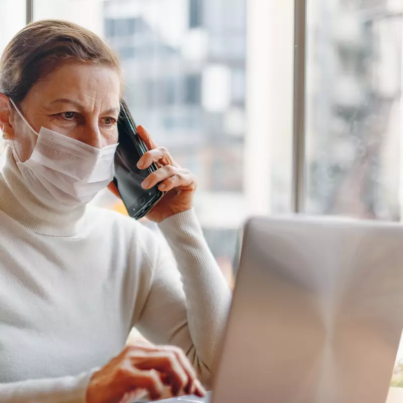 Mask wearing woman talking on the phone while using a laptop indoors.