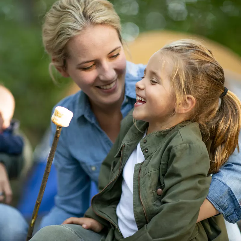 A family of four roasts marshmallows at a campground, the mother is holding her daughter as they smile.
