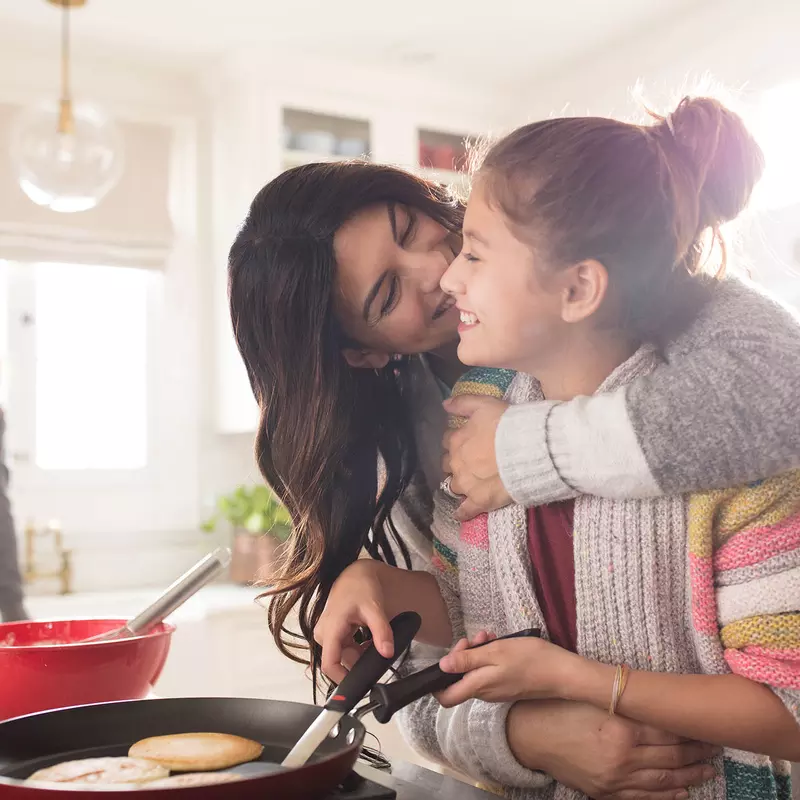 A family of three women are in the kitchen, one daughter hugging the youngest while she flips pancakes.