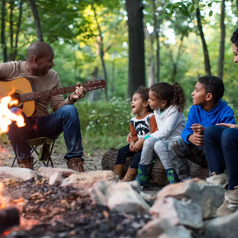 A family singing along while the father plays the guitar around the fireplace in the forest.