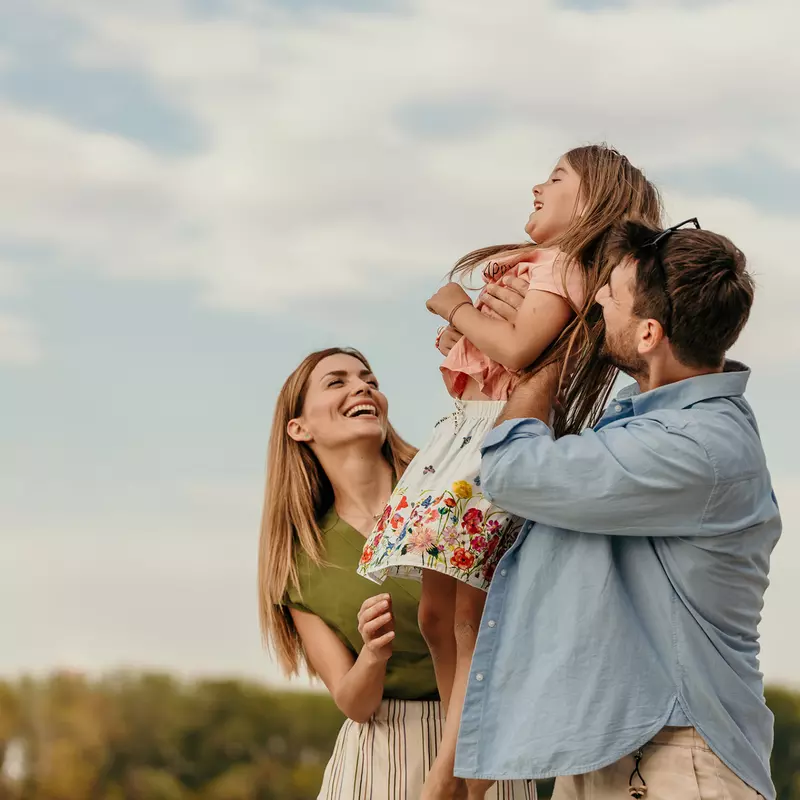 A family of a mother, father and daughter smiling while outdoors.