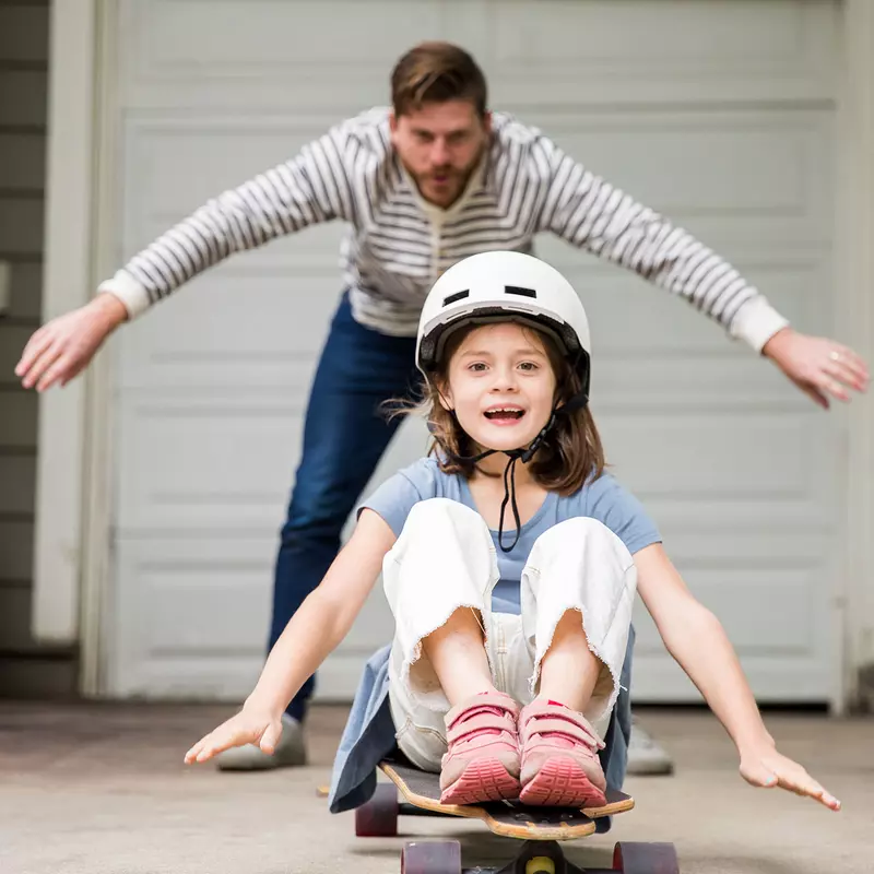 A dad showing his little girl how to skateboard in the carport.