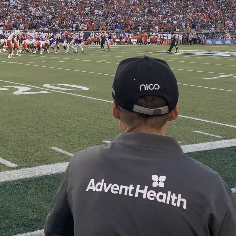 Man wearing shirt with AdventHealth logo on the sidelines of a football game.