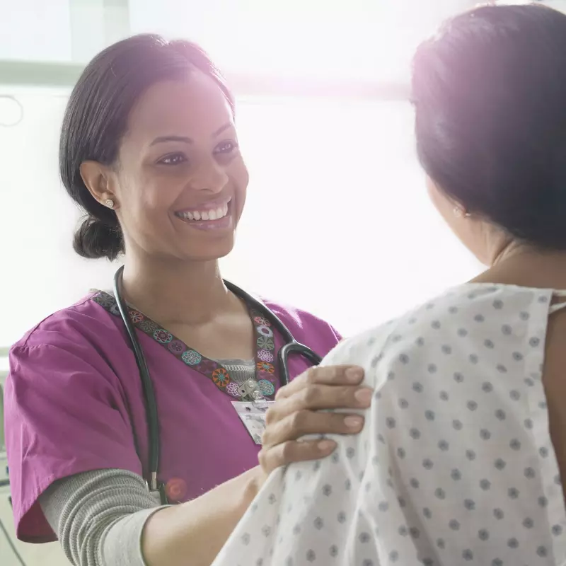 A smiling female physician reassures her female patient in an exam room.