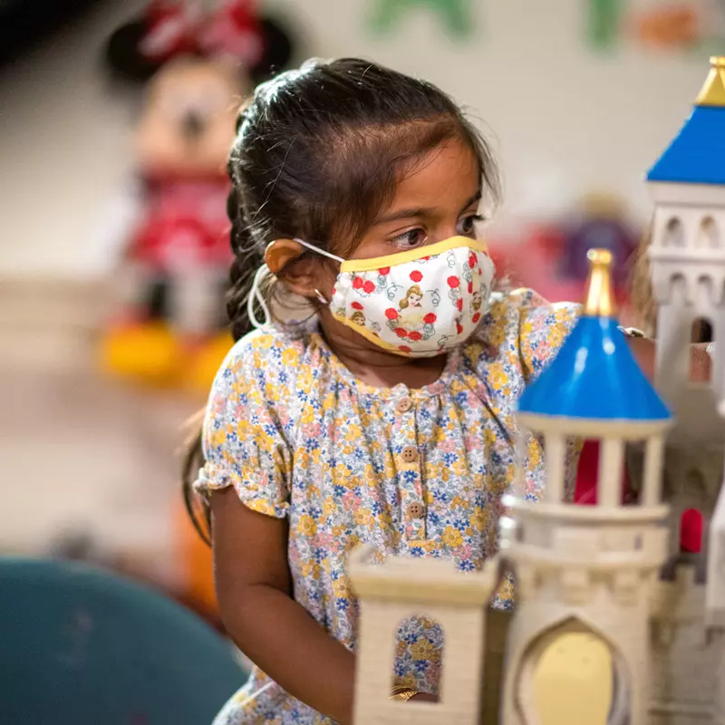 A little girl wearing a mask, playing with a Disney Castle play set. 