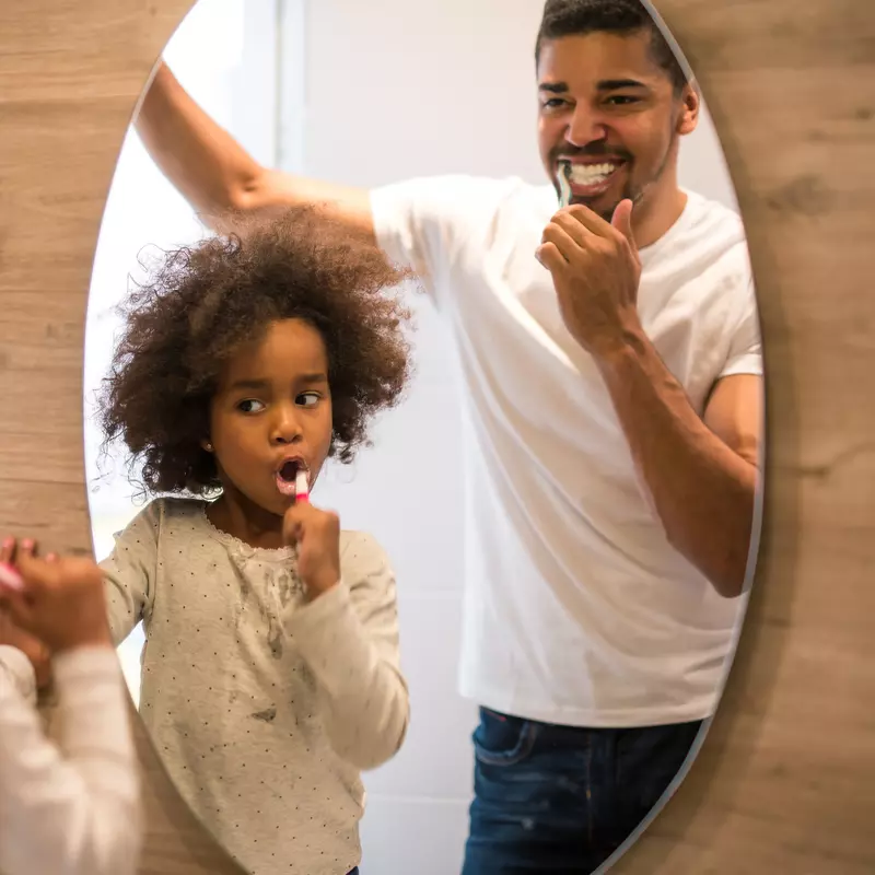 A father and daughter brush their teeth looking in a mirror.