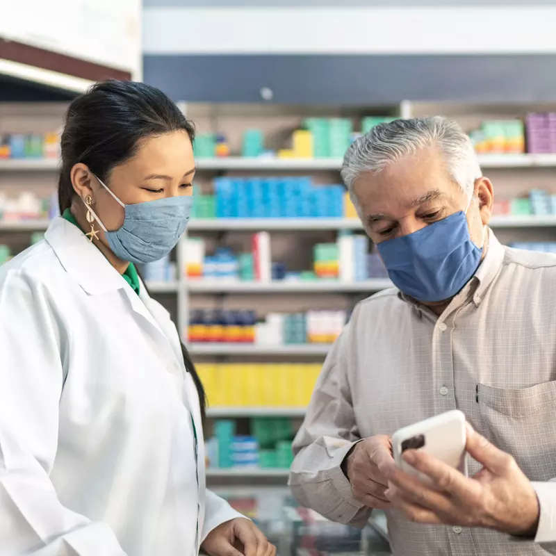 Older man talking with a pharmacist while both wear masks.