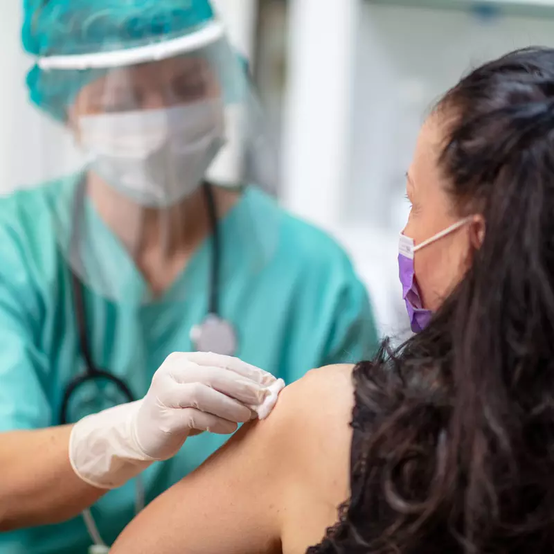 Provider prepping woman to receive a vaccine while they wear PPE and the woman wears a mask.