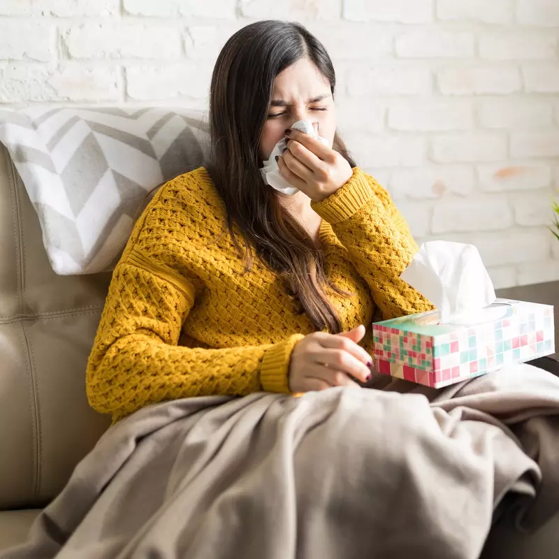 A young woman sitting on her couch sneezing. 