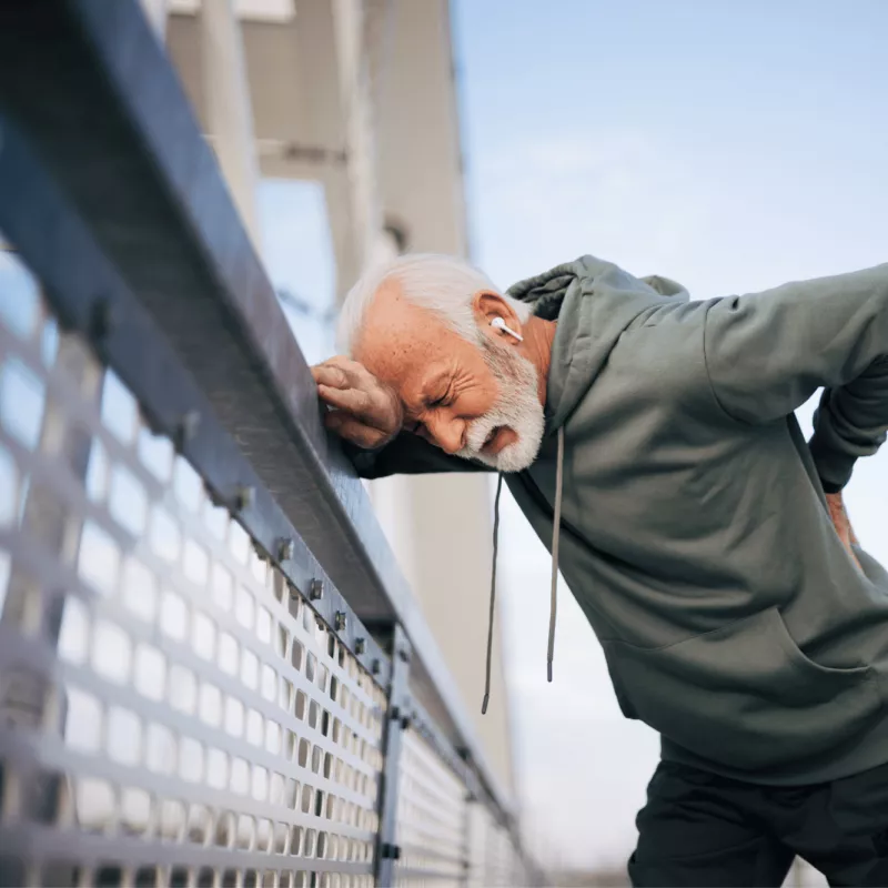 A senior man hunching over against a fence with his hand resting on his back in pain.