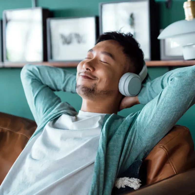 A man sits on a couch at home with listening to music with headphones with his eyes closed, smiling, and hands behind his head.