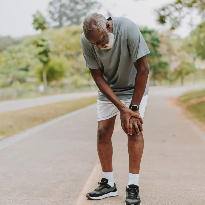 A senior black man takes a break from exercising outside and places his hands on his left knee.