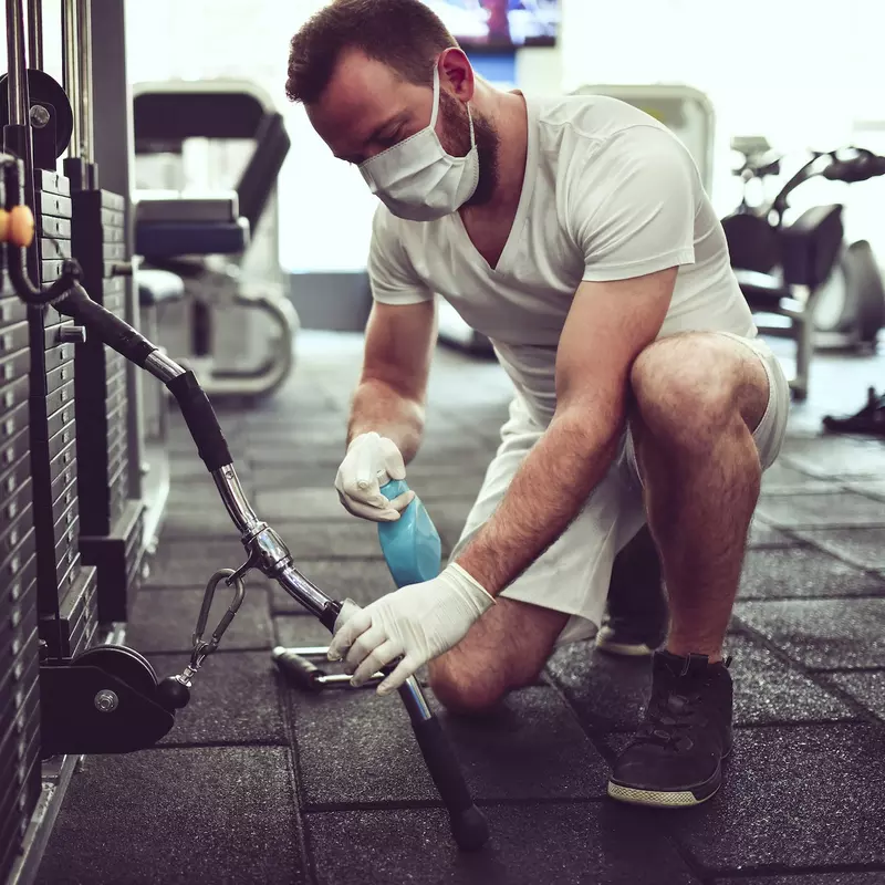 A man wearing a mask and cleaning fitness equipment in a gym.