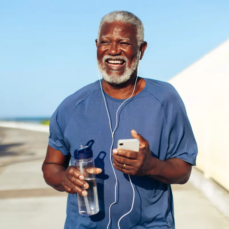 An older black man using headphones plugged into his phone while walking outdoors and holding a water bottle.