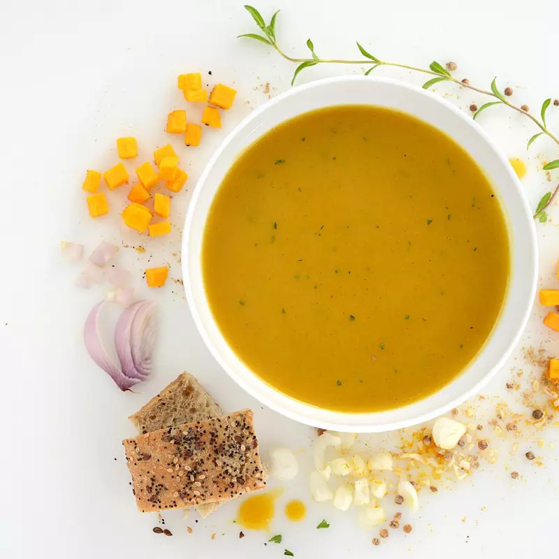 a bowl of creamy pumpkin soup, surrounded by herbs, spices, bread and chunks of pumpkin