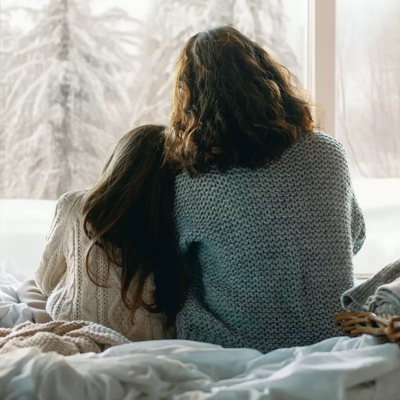 Mother and daughter looking through a window at the snow outside.