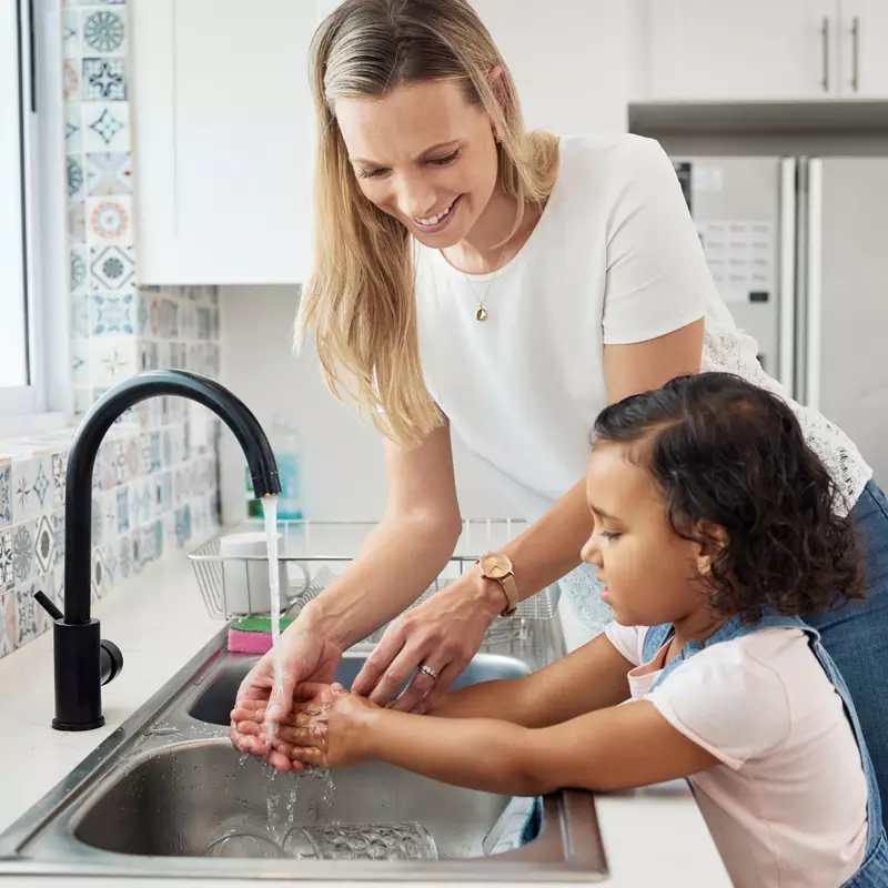 Mother showing her daughter how to wash her hands at the kitchen sink.