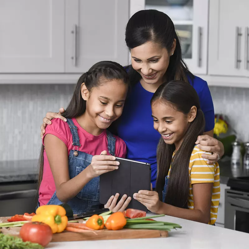 A mother and her 2 young daughters reading a recipe on a tablet in the kitchen.