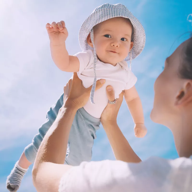 A mother holds up her baby whose wearing a hat while outside.