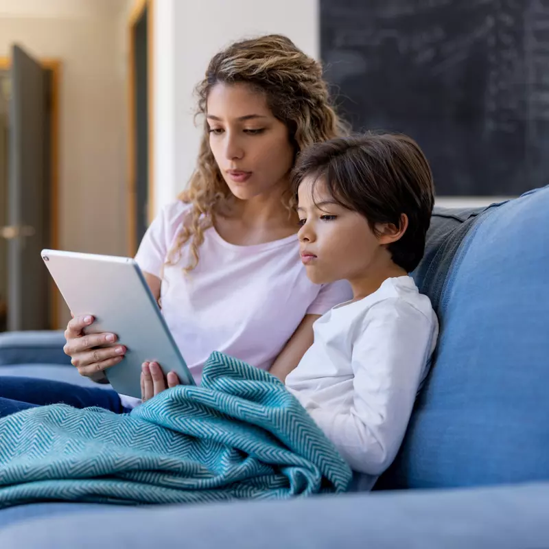 A mother and son sitting on a couch and using a tablet.