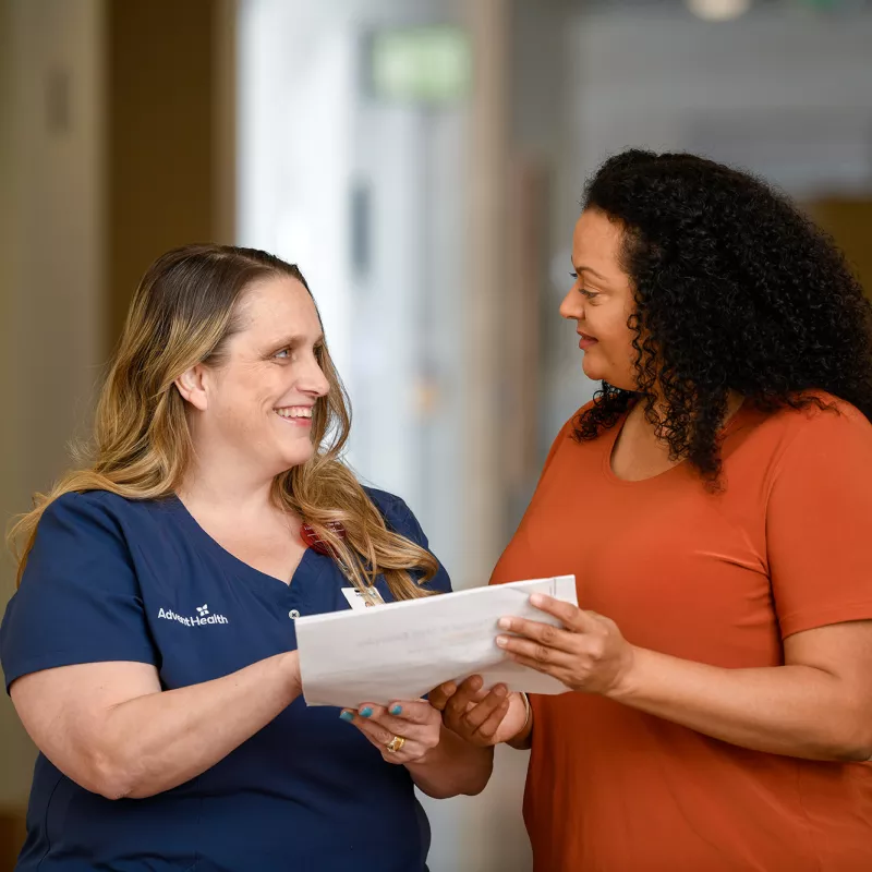 An AdventHealth nurse talking with a female patient while both hold a document.