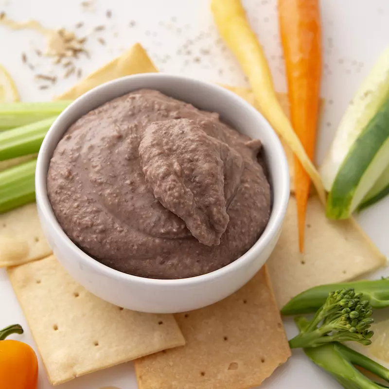 White bowl of black bean hummus surrounded by vegetable sticks and crackers.