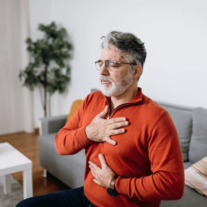 Senior man sitting on a couch at home placing hands on chest and abdomen.