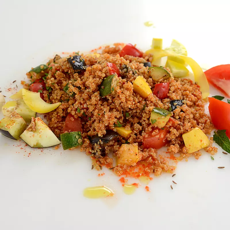 Mound of vegetables and quinoa on white surface with tomato garnish.