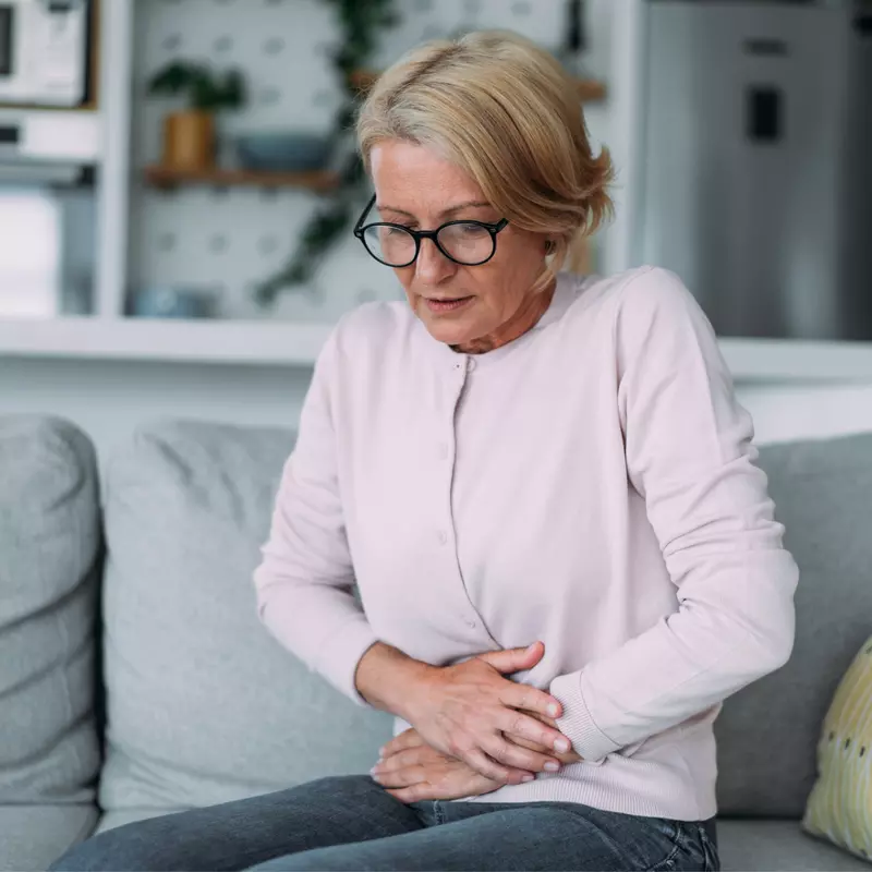 A senior woman sitting on a couch holding her stomach in discomfort while at home.