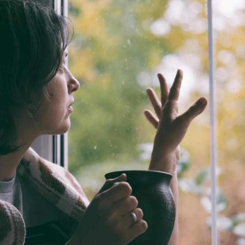 Woman looking out and touching window while indoors while wrapped in a blanket and holding a mug.