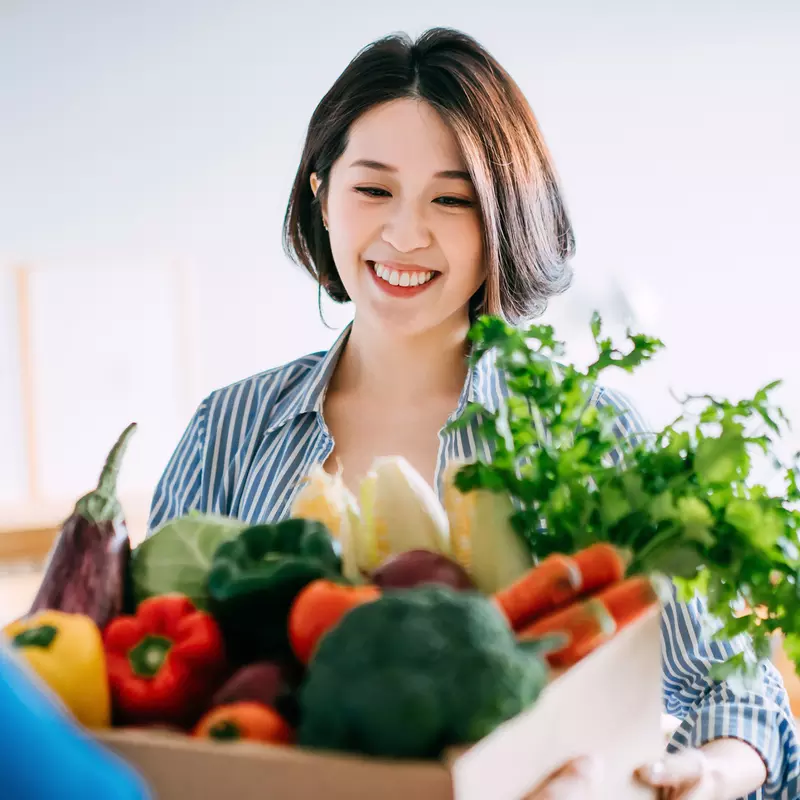 Woman receiving a box full of a variety of vegetables from another person.