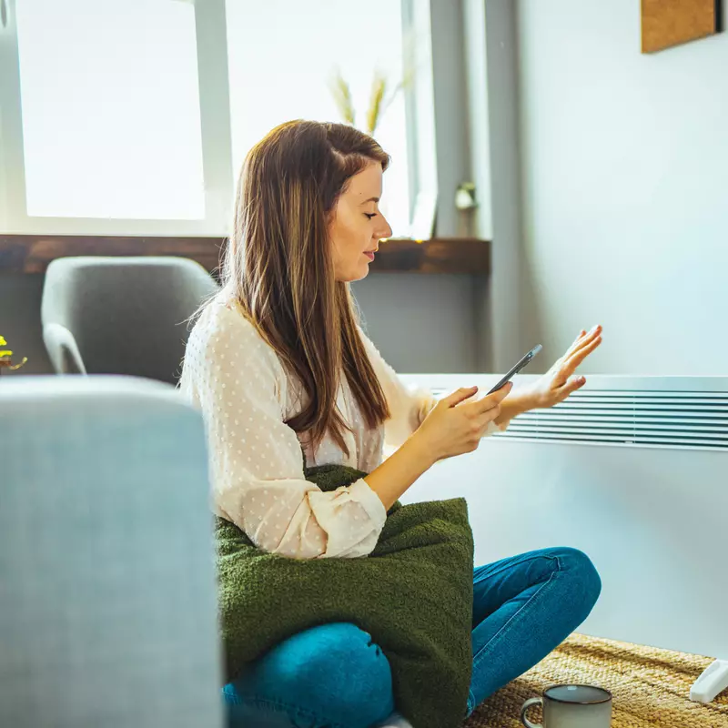 Woman using her phone while sitting next to a space heater at home.