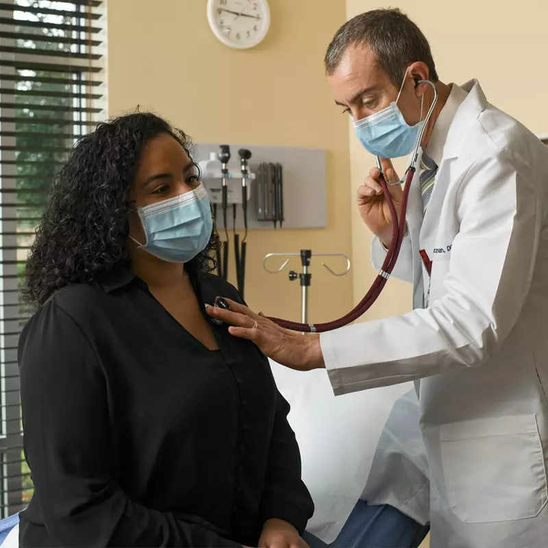 Doctor checking woman's heartbeat with stethoscope while both are wearing masks.